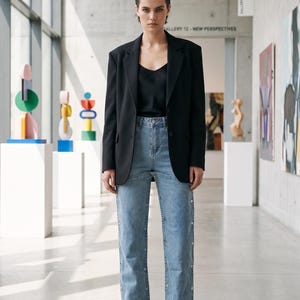 K&ouml;nnte beinhalten: Eine Person tr&auml;gt einen schwarzen Blazer &uuml;ber einem schwarzen Oberteil und hellblauen Jeans mit Kn&ouml;pfen. Dazu werden schwarze Loafer getragen. Der Hintergrund ist eine moderne Kunstgalerie mit farbenfrohen Skulpturen und Gem&auml;lden.