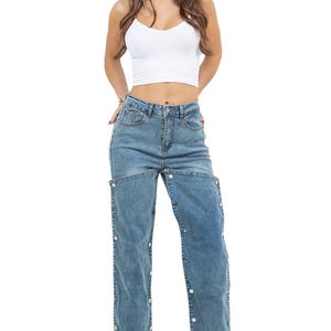 K&ouml;nnte beinhalten: Hellblaue Jeans mit Knopfdetails an den Au&szlig;enn&auml;hten. Die Jeans wird mit einem wei&szlig;en Crop-Top kombiniert. Das Model tr&auml;gt High Heels.