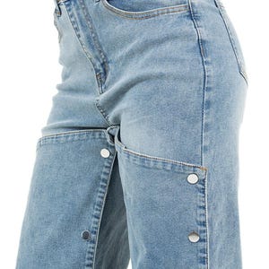 K&ouml;nnte beinhalten: Hellblaue Jeans mit einem einzigartigen Design. Die Jeans haben eine hohe Taille, klassisches F&uuml;nf-Taschen-Design und einen Knopfverschluss. Die Vorderseite der Jeans hat ein geteiltes Design mit silbernen Kn&ouml;pfen entlang der N&auml;hte.