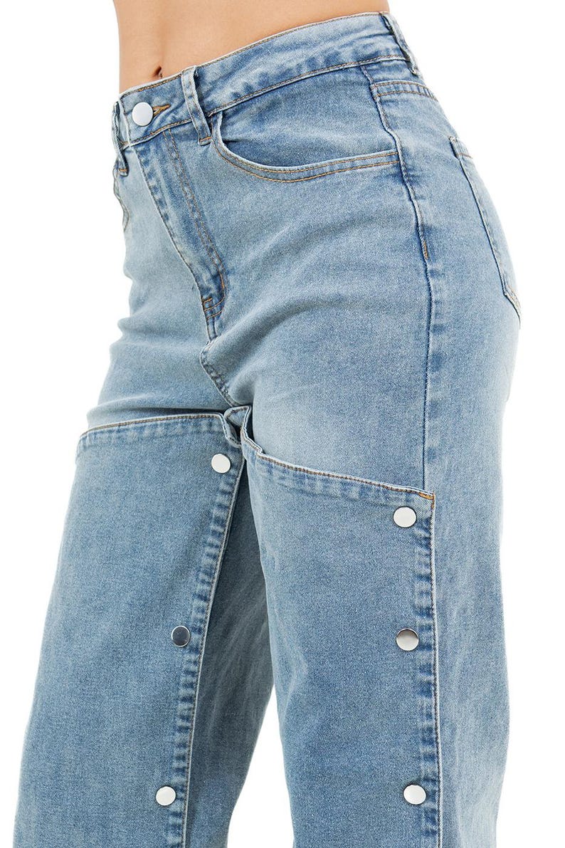 K&ouml;nnte beinhalten: Hellblaue Jeans mit einem einzigartigen Design. Die Jeans haben eine hohe Taille, klassisches F&uuml;nf-Taschen-Design und einen Knopfverschluss. Die Vorderseite der Jeans hat ein geteiltes Design mit silbernen Kn&ouml;pfen entlang der N&auml;hte.