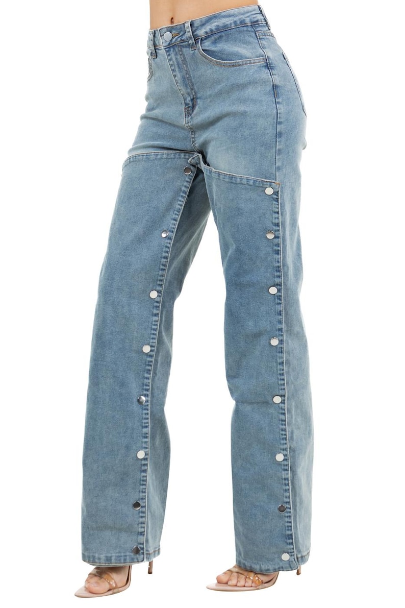 K&ouml;nnte beinhalten: Hellblaue Jeans mit einem einzigartigen Design. Die Jeans haben ein weites Bein und eine Reihe silberfarbener Kn&ouml;pfe entlang der Au&szlig;enn&auml;hte. Der Bund ist ein Standarddesign.
