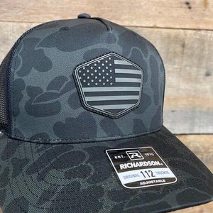 May include: A black and gray camouflage trucker hat with a mesh back. The hat features a gray patch with an American flag design. The brim has a curved shape, and the hat is adjustable. The Richardson brand logo is visible on the front.