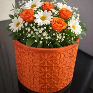 Könnte beinhalten: Ein orangefarbener Blumentopf gefüllt mit einem Blumenarrangement. Das Arrangement umfasst orangefarbene Rosen, weiße Gänseblümchen und kleine weiße Blüten. Der Topf hat ein strukturiertes, sich wiederholendes Muster.