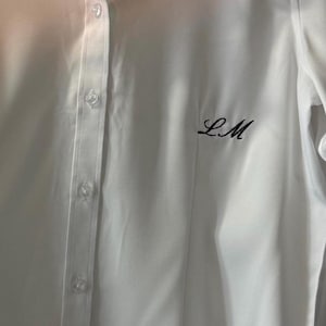 May include: A white button-down shirt with long sleeves. The shirt has a classic collar and button closure. The initials "LM" are embroidered in black thread on the left chest. The fabric appears smooth and slightly shiny.