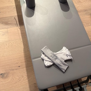 May include: A gray Pilates reformer with two gray headrests. A gray headband with embroidered text and a white ribbed wristband with ruffled edges are on the reformer. The reformer is on a wooden floor.