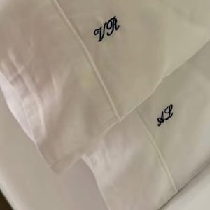 May include: Two white pillowcases with embroidered initials in dark blue thread. The top pillowcase displays "VR", and the bottom pillowcase shows "AL". The bedding appears to be made of a smooth, soft fabric.