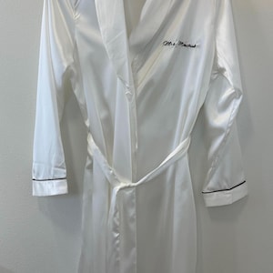 May include: White satin robe with a shawl collar and long sleeves. The robe has a matching belt and black trim on the cuffs. The text "Mrs. Minchuk" is embroidered on the chest. The robe is hanging on a black hanger.