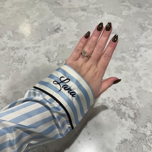 May include: A light blue and white striped pajama top sleeve with the name "Lana" embroidered in black script. The sleeve has a black trim. The hand has dark green nail polish and a gold ring.