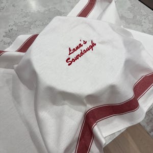 Op de afbeelding: Witte keukendoek met rode strepen en de geborduurde tekst "Lana's Sourdough". De doek is over een rond object, mogelijk brood, gevouwen en op een lichtgekleurd oppervlak geplaatst. De doek heeft een visgraatpatroon.