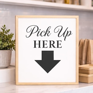 May include: A framed sign with the words "Pick Up HERE" in black script and a downward-pointing arrow. The sign is in a light wood frame and sits on a white countertop. A small potted plant and other cafe items are in the background.