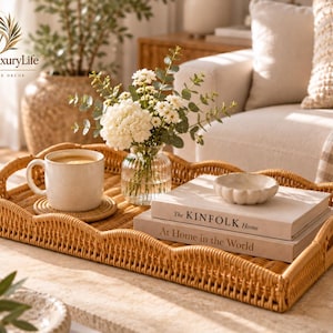 Puede incluir: Una bandeja rectangular de ratán trenzado con borde festoneado, que contiene una taza de cerámica sobre un posavasos, un jarrón de cristal con flores blancas, un pequeño cuenco festoneado y dos libros apilados titulados The Kinfolk Home y At Home in the World.