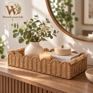 May include: A rectangular woven rattan tray with a scalloped edge and two light wood handles. Inside the tray sits a cream-colored ceramic vase with green foliage, a ribbed glass candle, and a book titled The KINFOLK Home with a box.