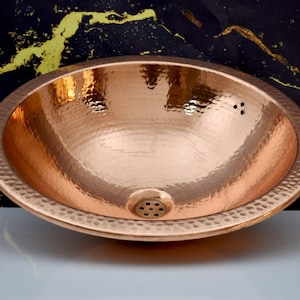 Può includere: Un lavabo ovale in rame martellato con una superficie strutturata e uno scarico. Il lavabo ha una finitura lucida e un bordo martellato decorativo. Il lavabo in rame è progettato per l'uso in bagno.