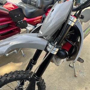 May include: A gray Kawasaki motorcycle with a black front tire and suspension. The motorcycle has a red air filter and a silver frame. A red motorcycle is partially visible in the background.