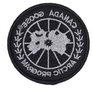 May include: A circular black embroidered patch featuring a white map of the Arctic region at the centre. The text CANADA GOOSE ARCTIC PROGRAM is stitched in white around the border, accompanied by white leaf-like symbols and a thin white grid.