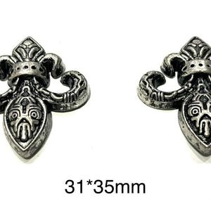 Op de afbeelding: Twee antieke zilverkleurige fleur-de-lis ornamenten. Elk ornament heeft een gedetailleerd ontwerp met een centrale schildvorm en gebogen accenten. De afmetingen zijn 3,1 cm bij 3,5 cm.
