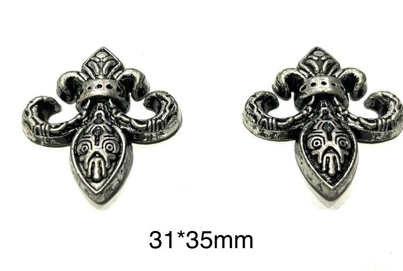 Op de afbeelding: Twee antieke zilverkleurige fleur-de-lis ornamenten. Elk ornament heeft een gedetailleerd ontwerp met een centrale schildvorm en gebogen accenten. De afmetingen zijn 3,1 cm bij 3,5 cm.