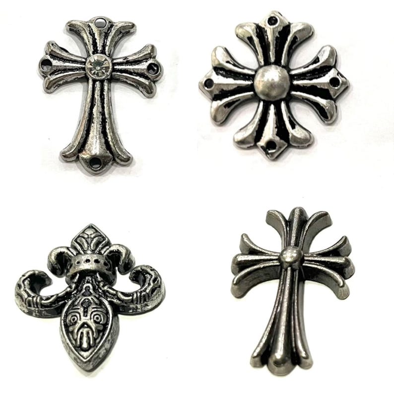 Op de afbeelding: Vier zilverkleurige metalen versieringen in verschillende ontwerpen. De ontwerpen omvatten twee kruisen, een fleur-de-lis en een cirkelvorm met een centrale stud. Elk stuk heeft een gedetailleerde, antieke afwerking.