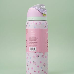 May include: A light pink Owala water bottle featuring a repeating pattern of small pink and blue flowers. The bottle has a light purple lid with a push-button mechanism and a lime green accent. A product information label is attached to the side.