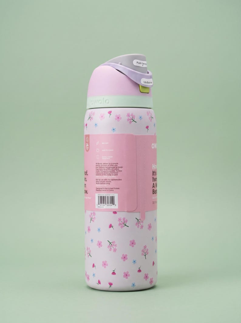 May include: A light pink Owala water bottle featuring a repeating pattern of small pink and blue flowers. The bottle has a light purple lid with a push-button mechanism and a lime green accent. A product information label is attached to the side.