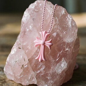 May include: A light pink cross pendant suspended on a matching pink ball chain. The cross features a smooth, matte finish with flared, rounded arms. The pendant is draped over a large, translucent, pale pink raw rose quartz crystal specimen.