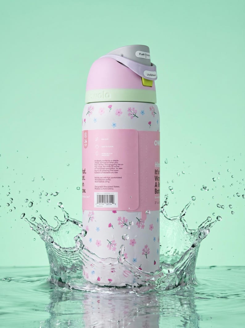 May include: A white Owala water bottle featuring a pink and blue floral pattern, a light green collar, and a grey lid with a push-button mechanism. The bottle has a pink product label wrapped around the centre with printed text and a barcode.