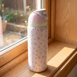 May include: A white Owala water bottle featuring a repeating pattern of small pink and blue flowers. The bottle has a light green collar and a grey lid with a push-button mechanism. The brand name Owala is printed in small letters on the collar.