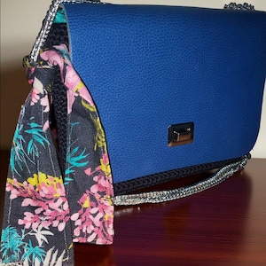 May include: A blue handbag with a silver chain strap and a decorative scarf. The scarf features a floral pattern with pink, yellow, and teal accents on a dark background. The bag has a silver clasp.