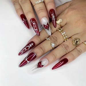 以下が含まれることがあります： 深紅のマニキュアとシルバーのアクセントが施された、長く尖った爪。一部の爪には複雑なシルバーのデザインが施され、他の爪にはハート型のデザインが施されています。宝石をあしらったいくつかの金の指輪が指を飾っています。