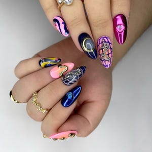 Puede incluir: Una mano con una variedad de uñas largas, ovaladas y manicuradas. Las uñas presentan diversos diseños, incluyendo patrones abstractos, acentos metálicos y adornos de gemas. Los colores incluyen rosa, azul, dorado y morado.