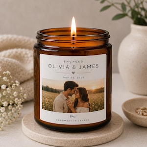 May include: An amber glass candle with a lit wick, featuring a label with the text "Engaged Olivia & James May 24, 2025." The label includes a photo of a couple. The candle is 227g and handmade in Canada.