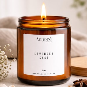 May include: A burning amber glass candle jar with a white label displaying "Lavender Sage" and "8 oz". The candle is from "Annoré Candle Studio" and is handmade in Canada. Decorative elements surround the candle, including a wooden lid.