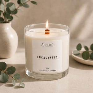 May include: A clear glass candle with a wooden wick and a white label reading Annoré Candle Studio, Eucalyptus, 8 oz, and Handmade in Canada. The candle contains white wax and is lit, emitting a small flame from the centre of the glass container.