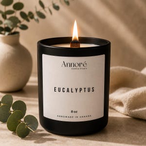 May include: A black candle in a matte container with a wooden wick and a lit flame. The white label reads Annoré Candle Studio, Eucalyptus, 8 oz, and Handmade in Canada. The candle is positioned upright with a visible wax surface.