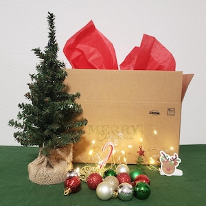 May include: A small artificial Christmas tree in a burlap base next to a brown cardboard box with red tissue paper. The box features the text Merry Christmas. A collection of red, green, gold, and silver ornaments, a candy cane, and fairy lights.