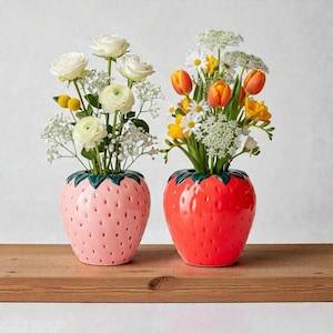 Könnte beinhalten: Zwei keramische Erdbeer-Vasen, eine rosa und eine rote, jeweils mit Blumen gefüllt. Die rosa Vase enthält weiße Rosen und Schleierkraut. Die rote Vase enthält orangefarbene Tulpen, Gänseblümchen und gelbe Blumen. Beide Vasen stehen auf einer Holzoberfläche.