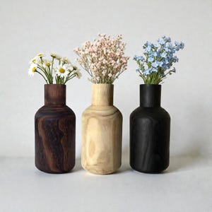 May include: Three wooden vases in varying shades of brown, beige, and black, each holding a different type of flower. The vase on the left is dark brown, the middle vase is light beige, and the right vase is black. The flowers include white daisies, pink baby's breath, and blue forget-me-nots.