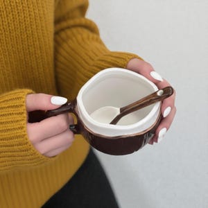 Puede incluir: Una taza de cerámica marrón y blanca con una cuchara a juego. La taza tiene un exterior texturizado y un interior blanco. La cuchara es de madera. La taza la sostiene una persona que lleva un jersey amarillo.