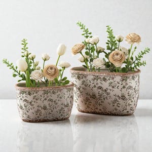 Può includere: Due fioriere ovali in ceramica con motivi floreali. Sono piene di fiori e verde bianchi, crema e beige. Le fioriere hanno uno sfondo beige chiaro con un disegno floreale marrone.