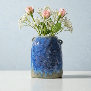 May include: A blue ceramic vase with a textured surface, holding pink roses and white baby's breath. The vase has a two-tone effect, transitioning from a deep blue to a lighter, earthy tone at the base. Small handles are on either side of the vase.