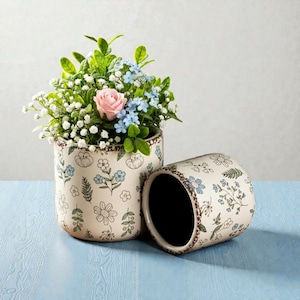 Könnte beinhalten: Zwei cremefarbene Keramik-Blumentöpfe mit floralem Design. Ein Topf steht aufrecht und ist mit einem Blumenstrauß gefüllt, darunter eine rosa Rose, blaue Vergissmeinnicht und weißes Schleierkraut. Der andere Topf liegt auf der Seite.