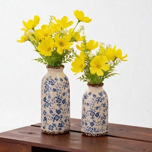 Op de afbeelding: Twee keramische vazen met blauwe bloempatronen bevatten heldergele bloemen. De vazen hebben een crèmekleurige achtergrond met blauwe bloemen en een bruine rand. De vazen staan op een houten oppervlak.