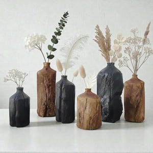Könnte beinhalten: Eine Sammlung rustikaler Holzvasen in verschiedenen Größen und Farben, darunter Braun und Schwarz. Jede Vase enthält getrocknete Blumen und dekorative Stiele, wodurch eine natürliche und elegante Präsentation entsteht. Die Vasen sind auf einer weißen Oberfläche angeordnet.