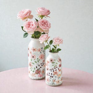 Peut inclure: Deux vases en céramique blanche à motifs floraux, contenant des roses roses. Les vases ont une finition texturée et vieillie et sont décorés de fleurs roses et marron et de feuilles vertes. Les vases sont sur une surface rose.
