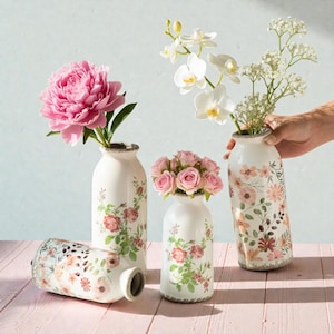 Op de afbeelding: Witte keramische vazen met bloemmotieven, met roze en witte bloemen. Eén vaas ligt op zijn kant. De vazen hebben een distressed rand en staan op een roze houten ondergrond.