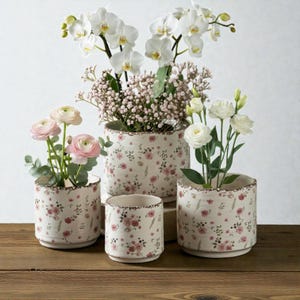 Könnte beinhalten: Vier Keramik-Blumentöpfe mit cremefarbenem Hintergrund und einem zarten rosa Blumenmuster. Die Töpfe, gefüllt mit Orchideen, Schleierkraut und Rosen, haben ein rustikales, gealtertes Aussehen. Unterschiedliche Größen.
