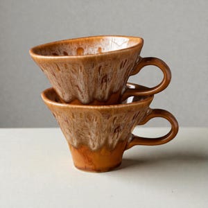 Könnte beinhalten: Zwei gestapelte braune Keramik-Kaffeetassen mit strukturierter, getropfter Glasur. Die Tassen haben eine einzigartige dreieckige Form und gebogene Griffe. Die Farbe ist ein warmes, erdiges Braun mit helleren Streifen.