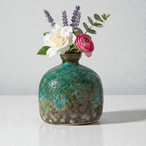 Könnte beinhalten: Eine kleine, runde Keramikvase mit einer strukturierten, türkis-braunen Glasur. Die Vase enthält einen Strauß frischer Blumen, darunter eine weiße Gardenie, ein rosa Ranunkel und Zweige von Lavendel und Eukalyptus. Die Vase steht auf einer weißen Oberfläche.