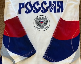 May include: White hockey jersey with red and blue accents. The word "РОССИЯ" in blue Cyrillic script is across the chest, above a circular emblem with a double-headed eagle and hockey sticks. The Nike logo is visible.