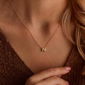 May include: A gold-coloured necklace featuring a small, polished letter N pendant suspended from a delicate, thin chain. The pendant has a smooth, rounded surface and sits centred on the chain, which rests against the skin of the wearer.